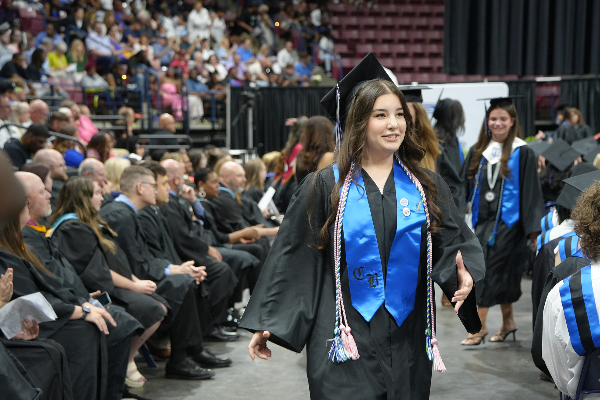 A woman in a blue and white graduation gown walks past seated graduates in an auditorium.