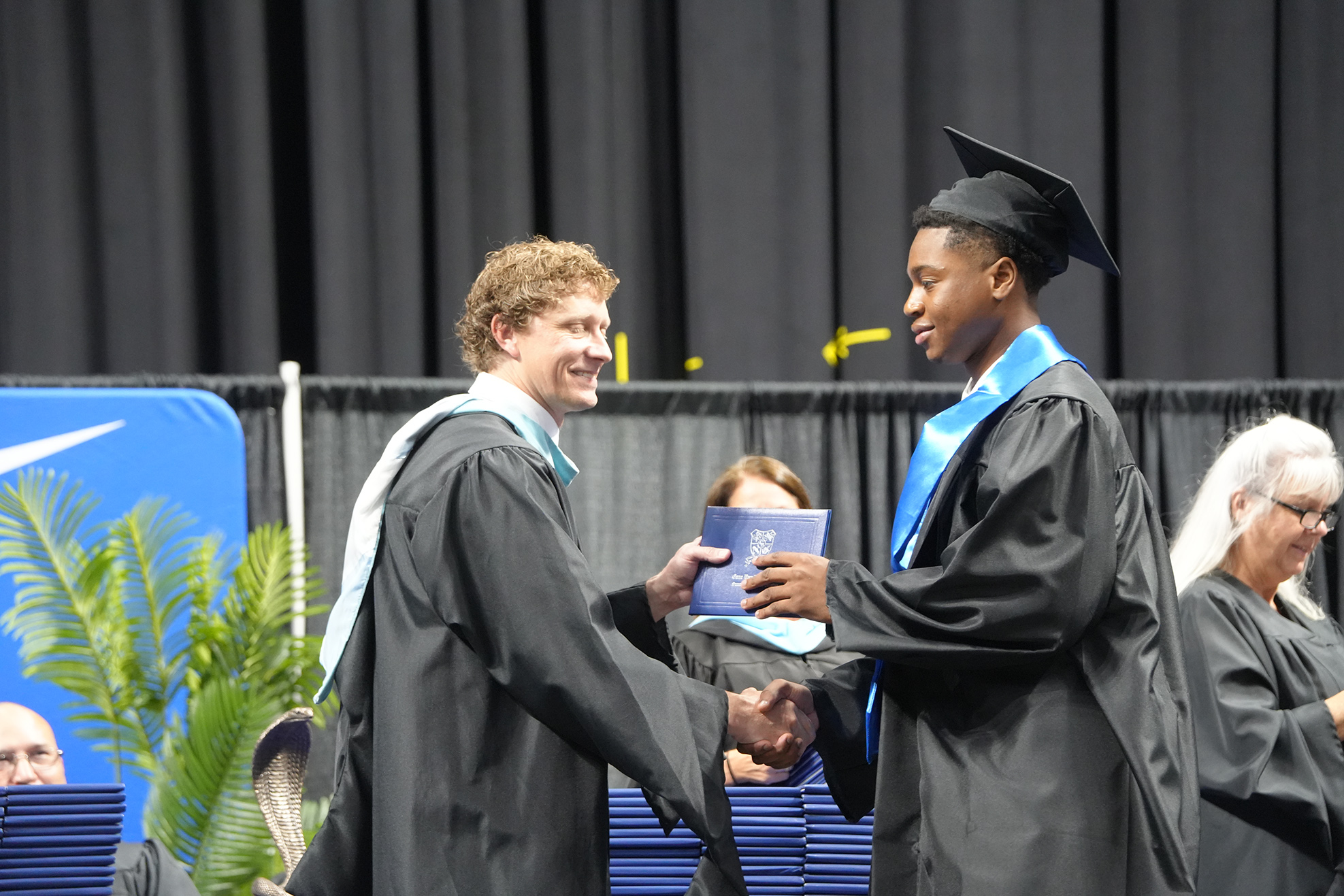 Two men in graduation gowns and caps exchange a document. A woman in the background watches.