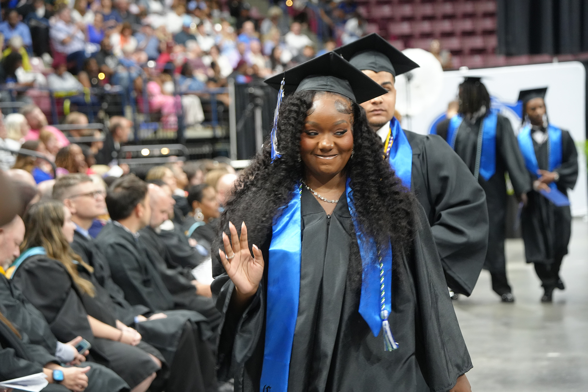 Woman in black graduation gown waves while walking in front of seated audience in black gowns.