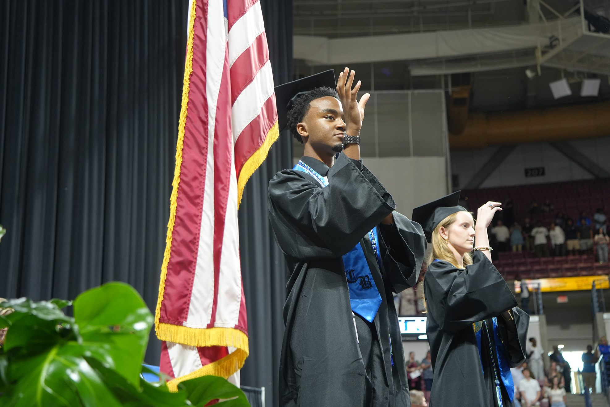 A man and a woman in academic regalia stand with hands raised. The man stands beside an American flag.