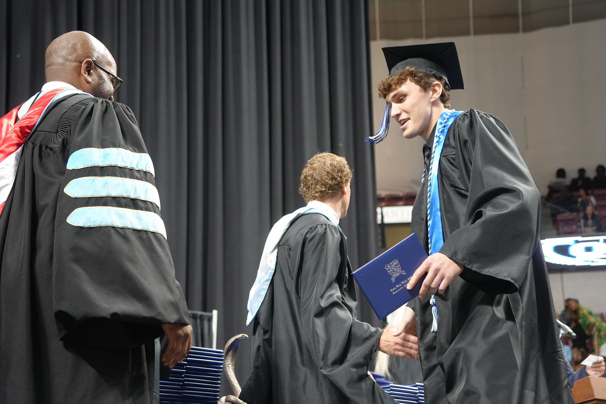 Three people in black graduation robes, one holding a blue diploma, stand on a stage.