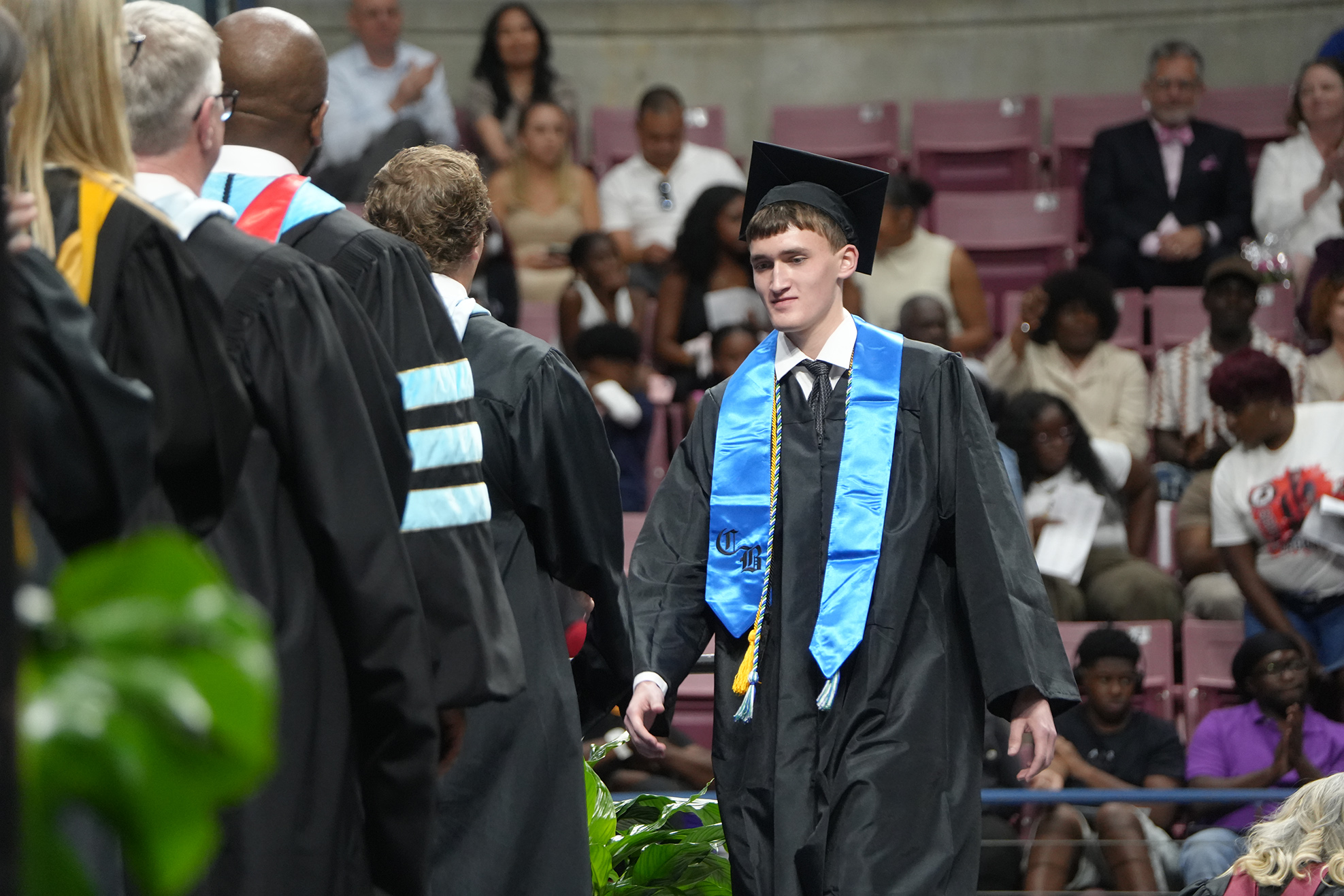 People in academic regalia walking in a line, with an audience seated in the stands.