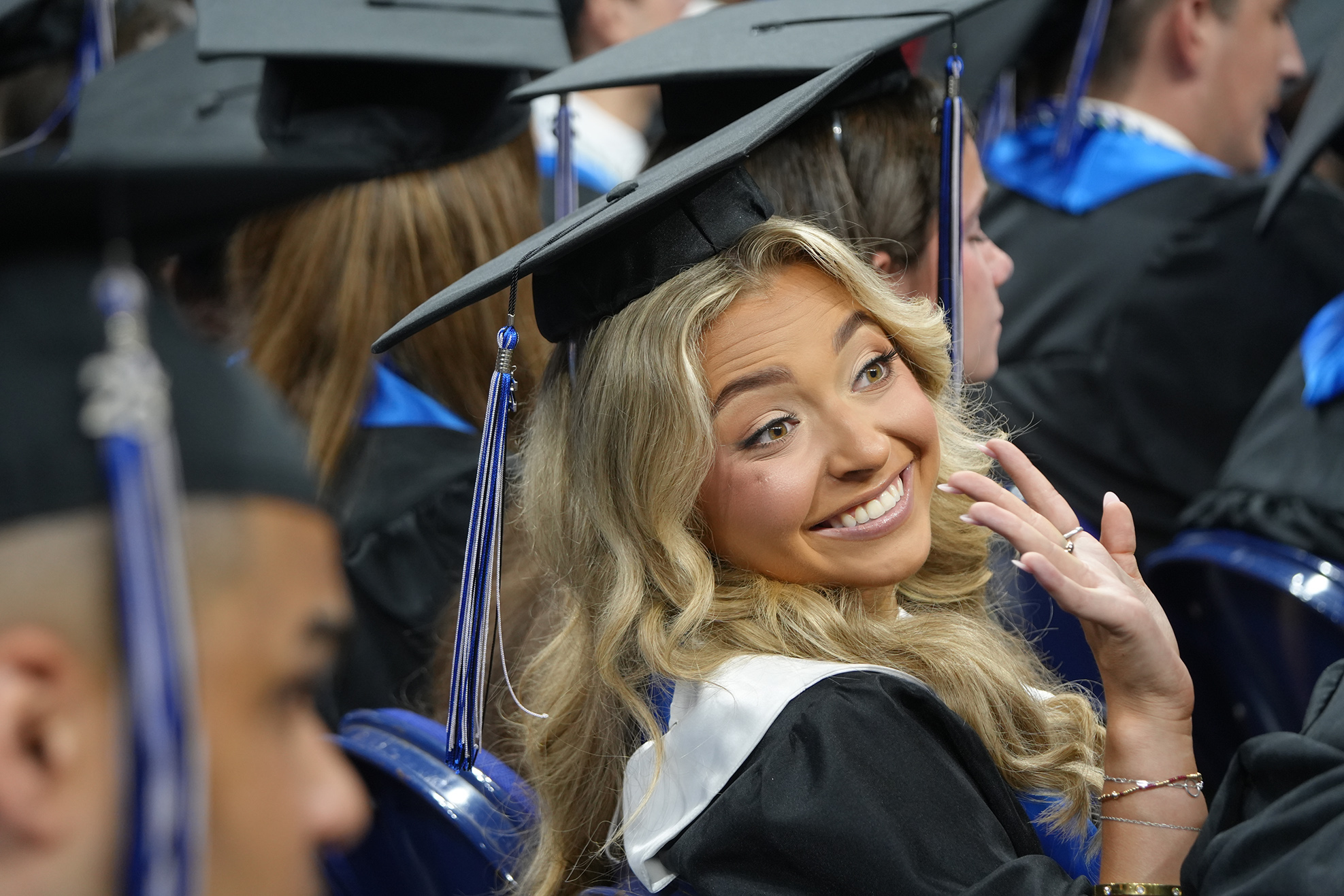 A woman in a cap and gown smiles and gestures, surrounded by others in similar attire at an event.
