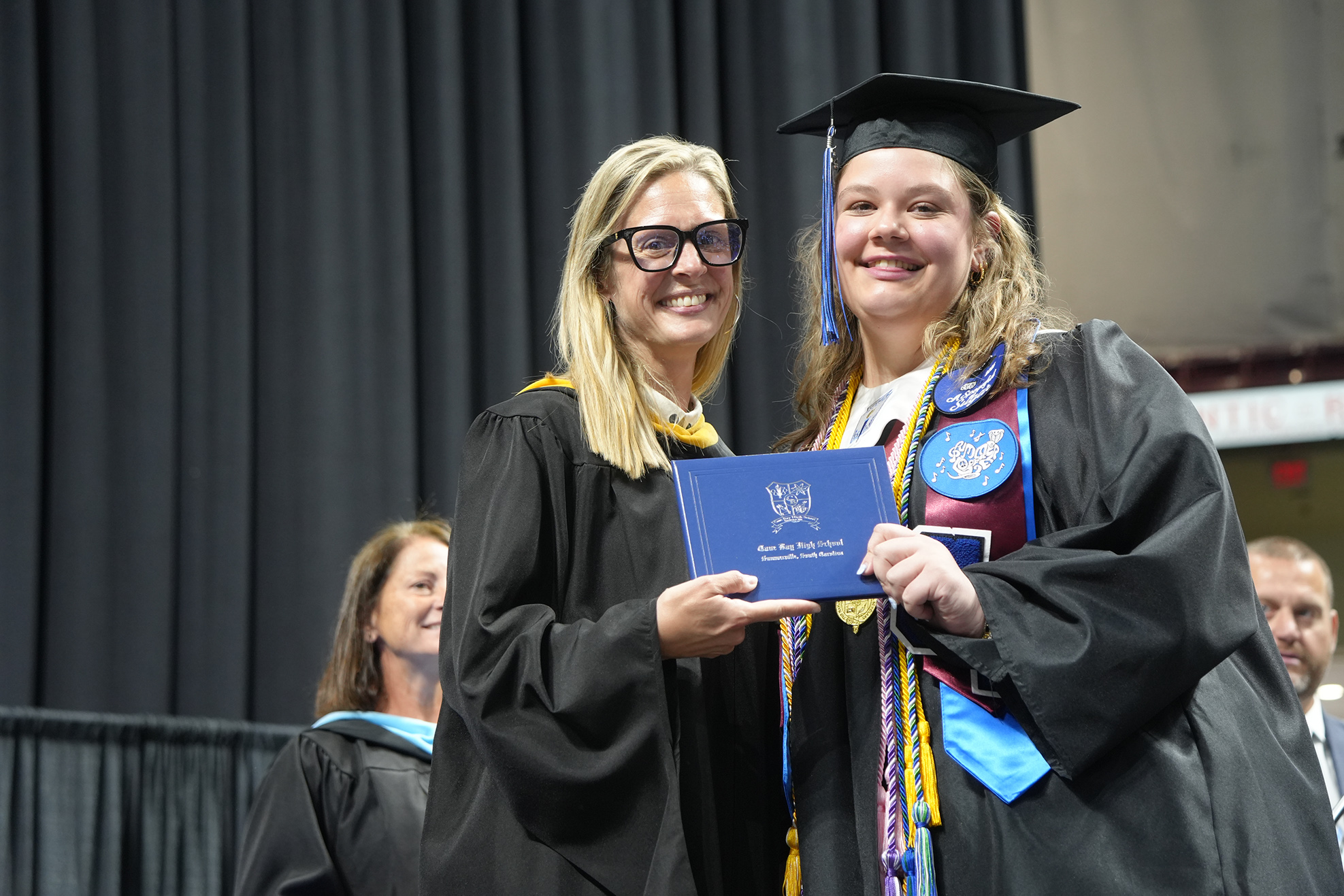 Two women in graduation attire on stage. One holds a blue diploma, the other holds it up. A blurred background features a black curtain.