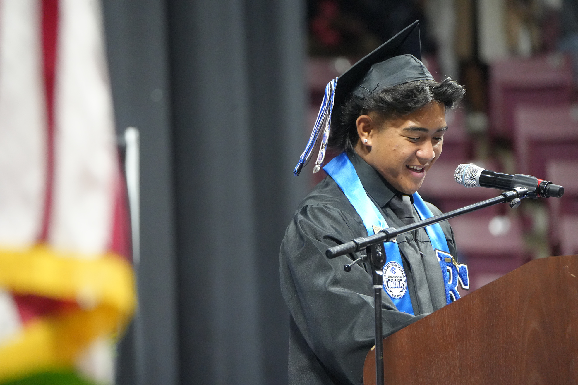 A person stands at a podium, speaking into a microphone, wearing a graduation cap and gown.