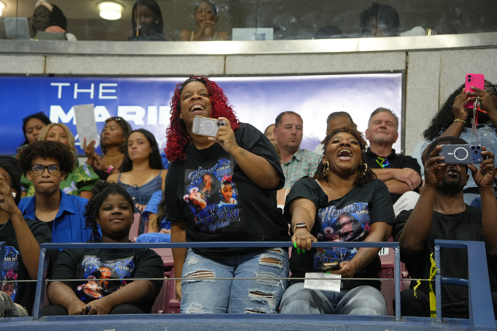 A group of people in an audience, some wearing black t-shirts, are cheering and holding up their phones.