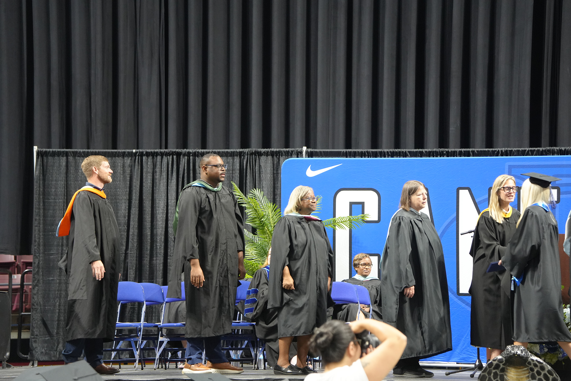 People in graduation gowns stand on a stage, possibly at a graduation ceremony, with a blue backdrop.