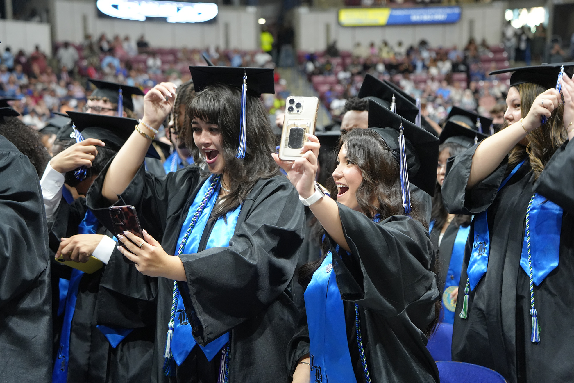 Graduates in black gowns and caps, some holding phones, stand in a stadium with a cheering crowd.