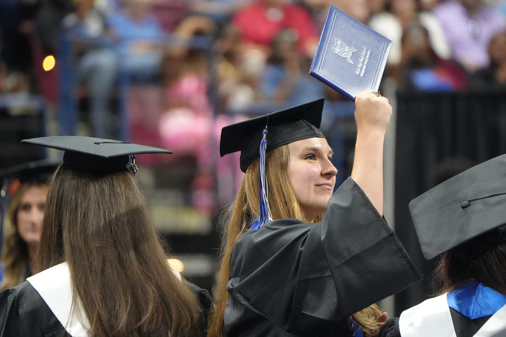 A female graduate holds a blue diploma in one hand and raises her other hand in front of others.