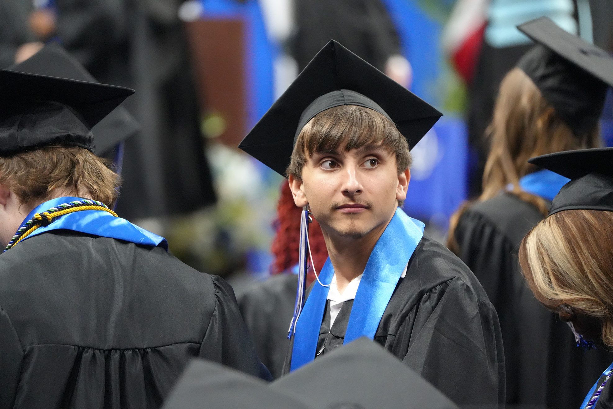 Graduates in black gowns and caps, one looking towards the front, stand among others at a ceremony.