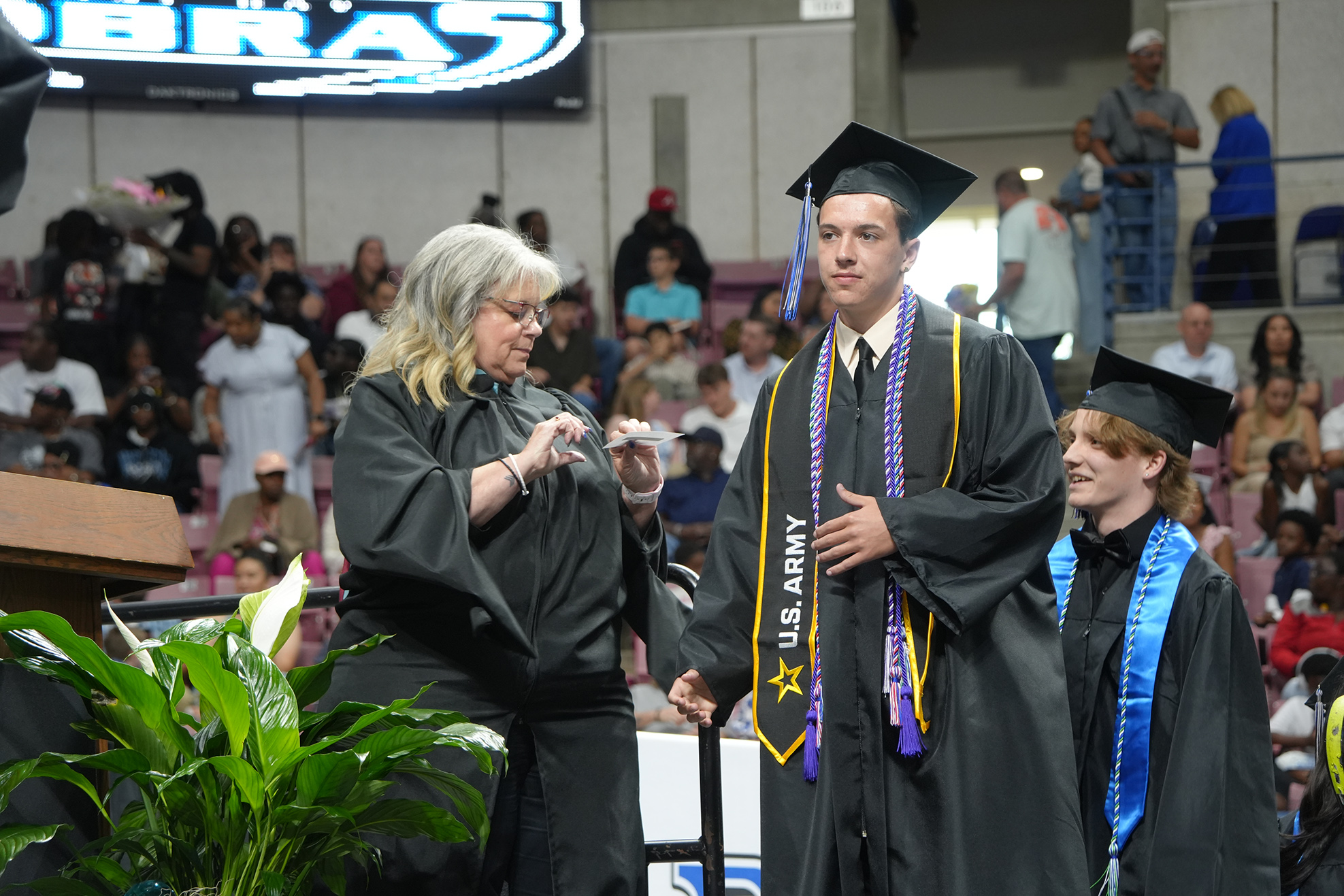 Two men in graduation robes, one with a sash, stand next to a woman at a ceremony.