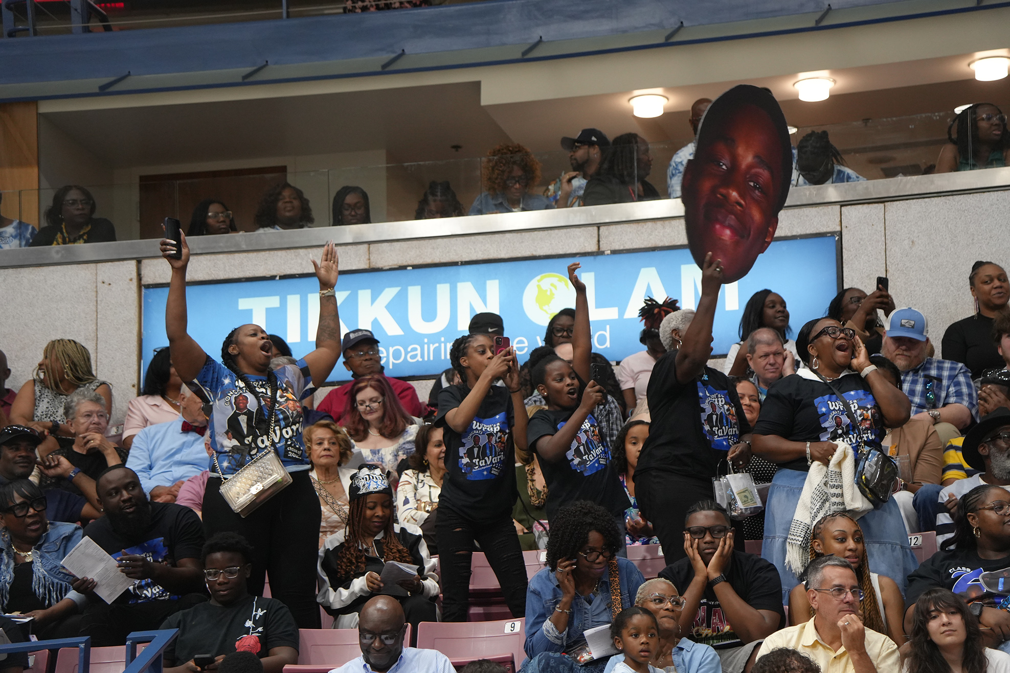 Spectators in a stadium stand with hands raised, holding a cardboard cutout of a smiling face.