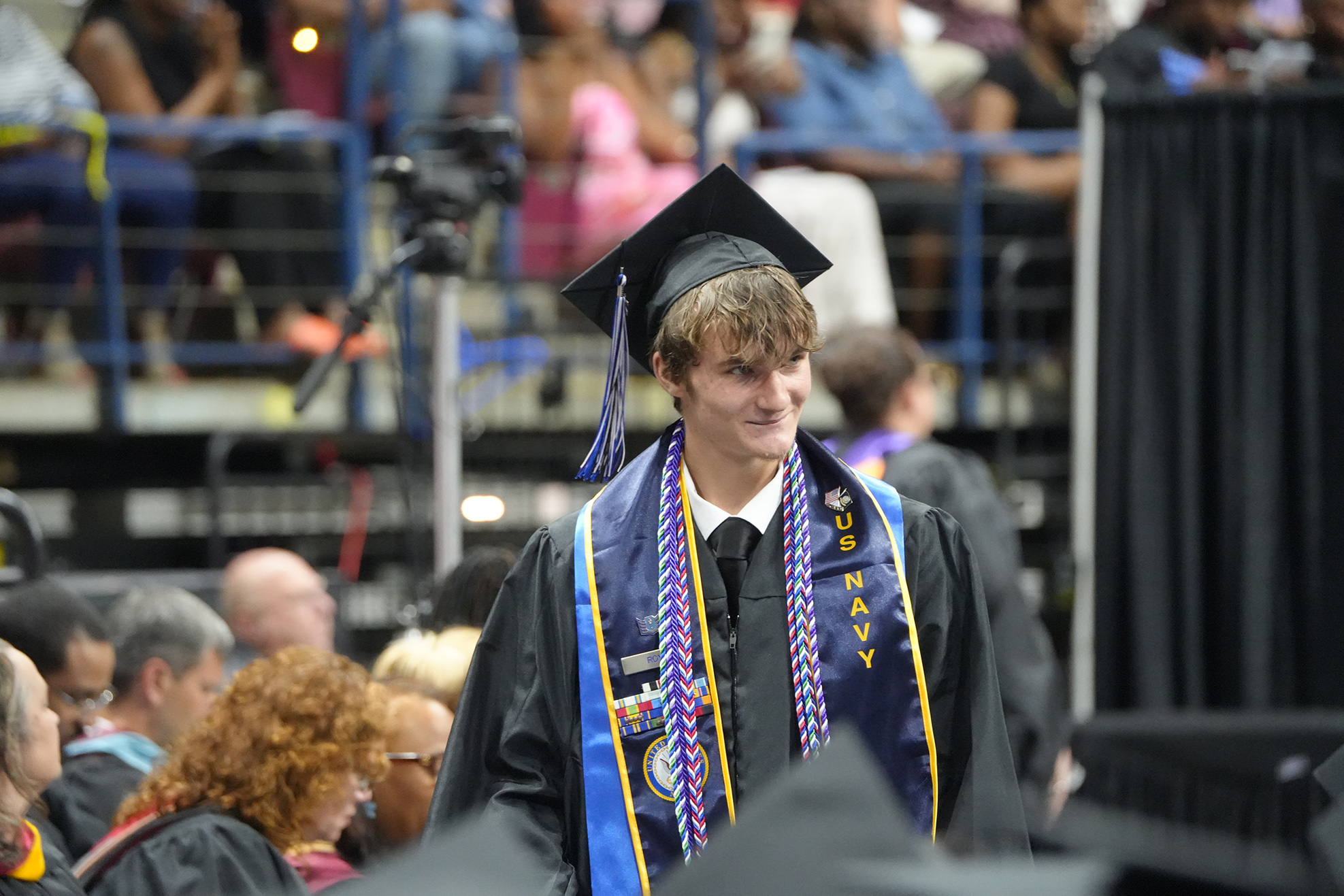 A man in a graduation gown with a blue sash walks past a crowd of people in an auditorium.