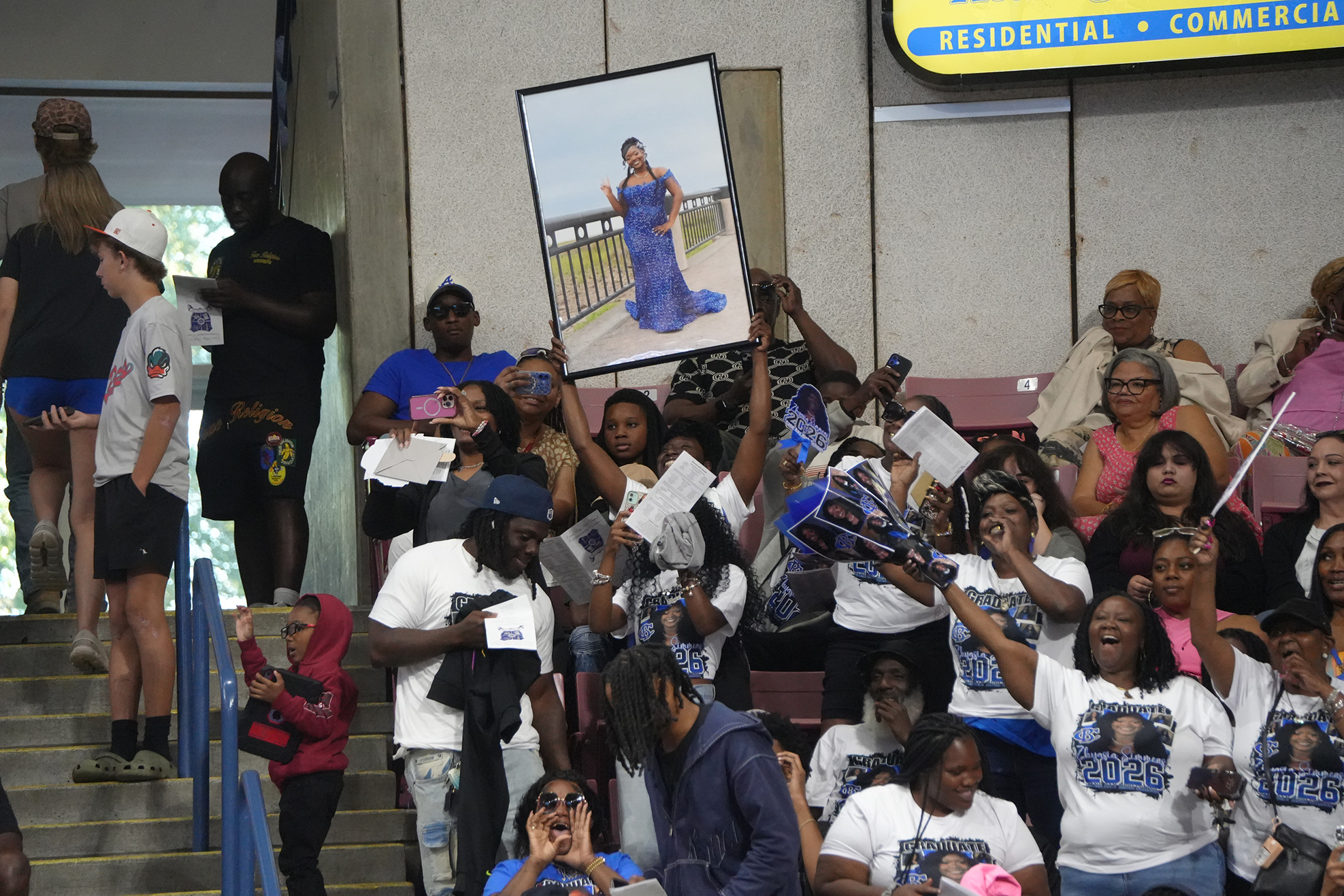 Crowd in stands at a sporting event. Some holding signs, others cheering. Some wearing matching white T-shirts.