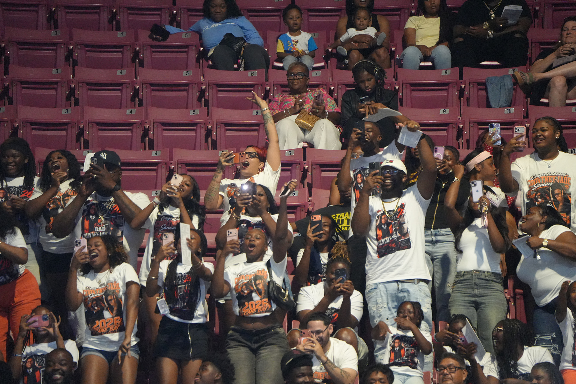Spectators in a stadium, many wearing matching white T-shirts, are cheering and raising their arms.