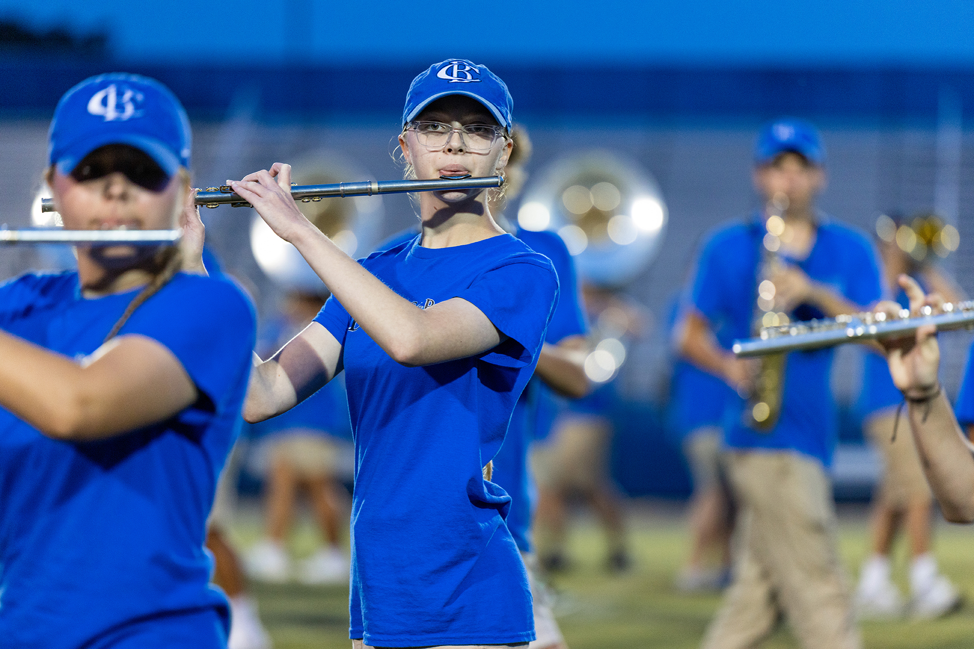 A band performs on a field. Two people play flutes. Two others play saxophones. The band wears blue shirts and pants.