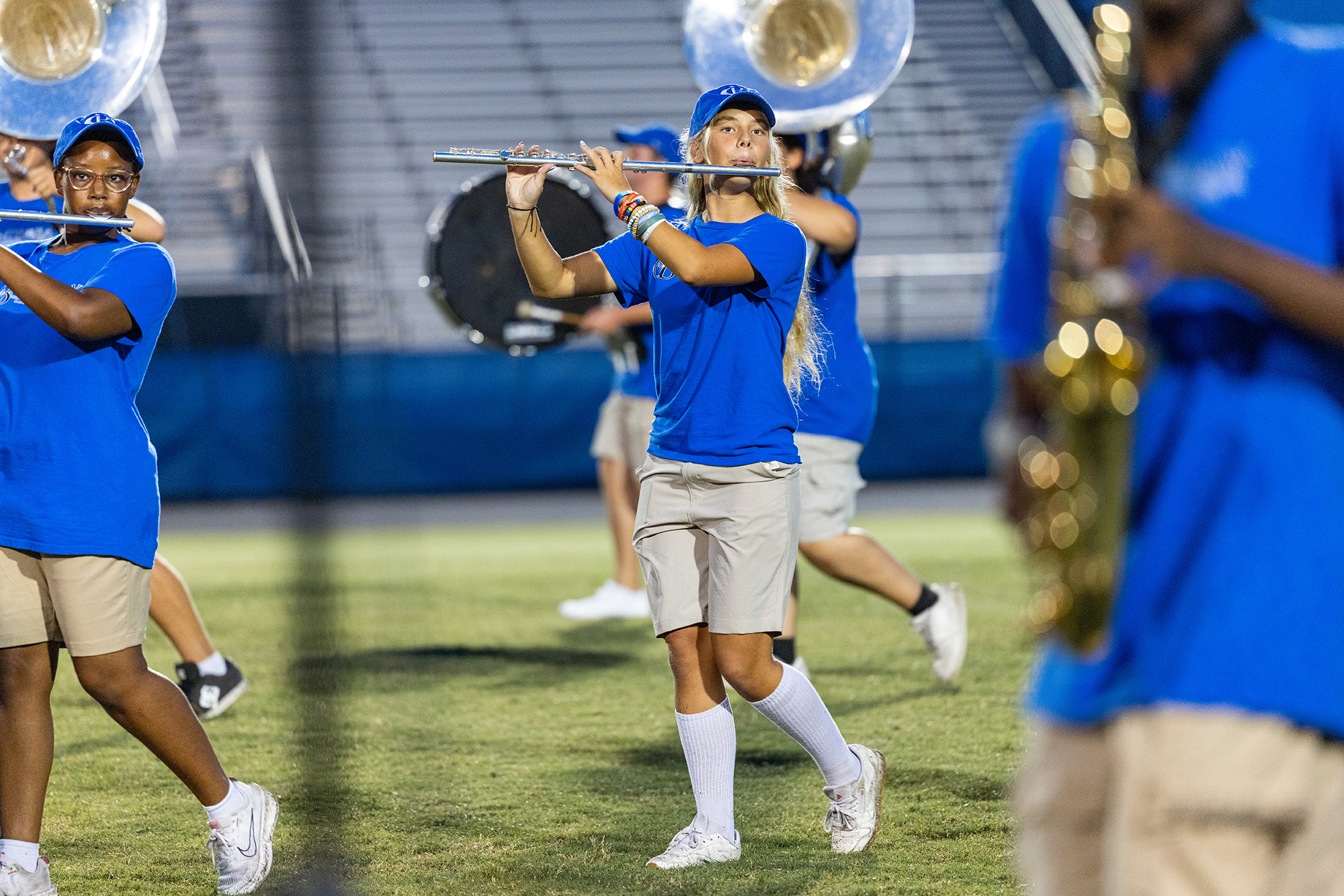 Group of young adults in blue shirts and khaki shorts play instruments on a grassy field.