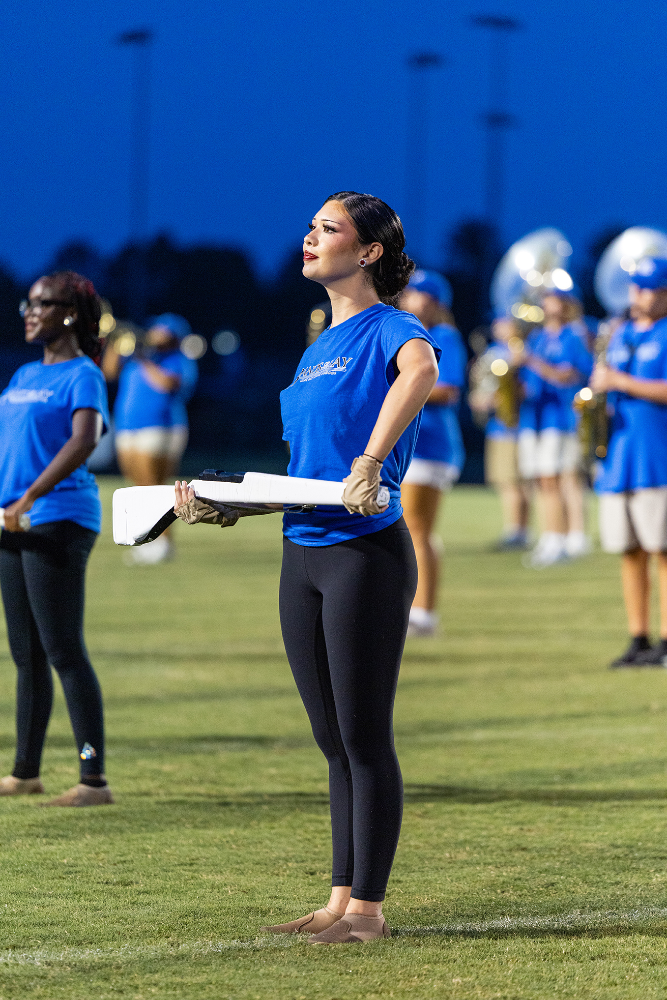 A woman in a blue top and black leggings stands on a grassy field, holding a baton. Other individuals in similar attire are in the background.