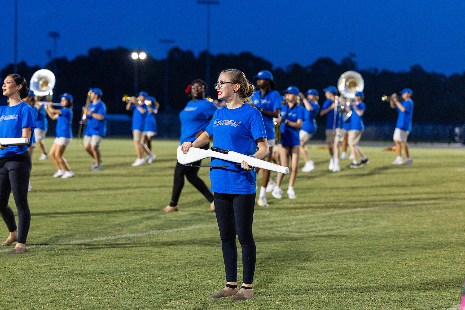 A marching band practices on a field at night. The musicians hold instruments. The field has lines and trees in the distance.