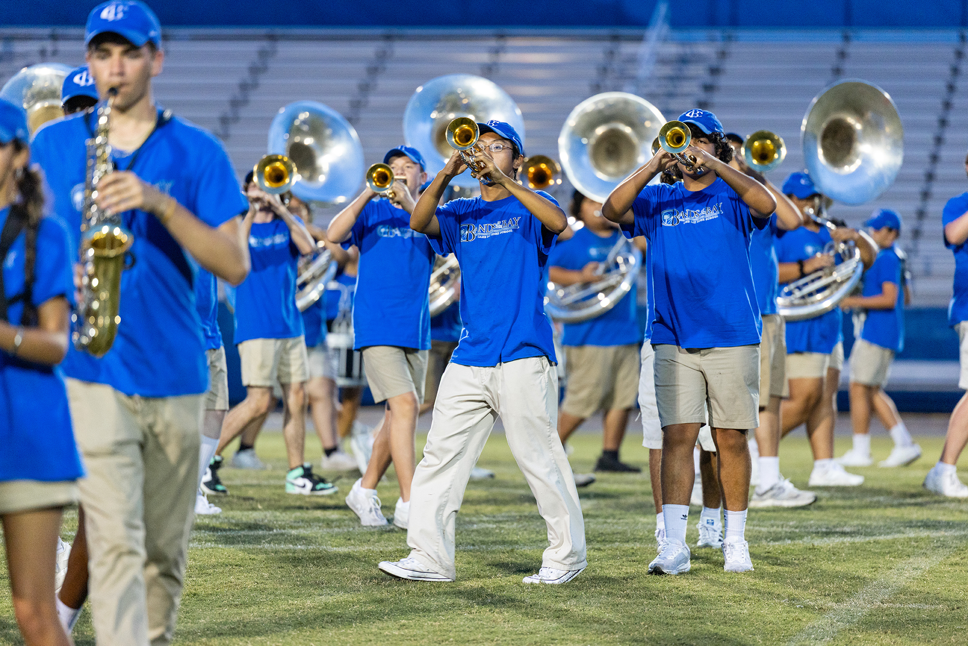 A group of musicians in blue uniforms and khaki shorts play instruments on a field.