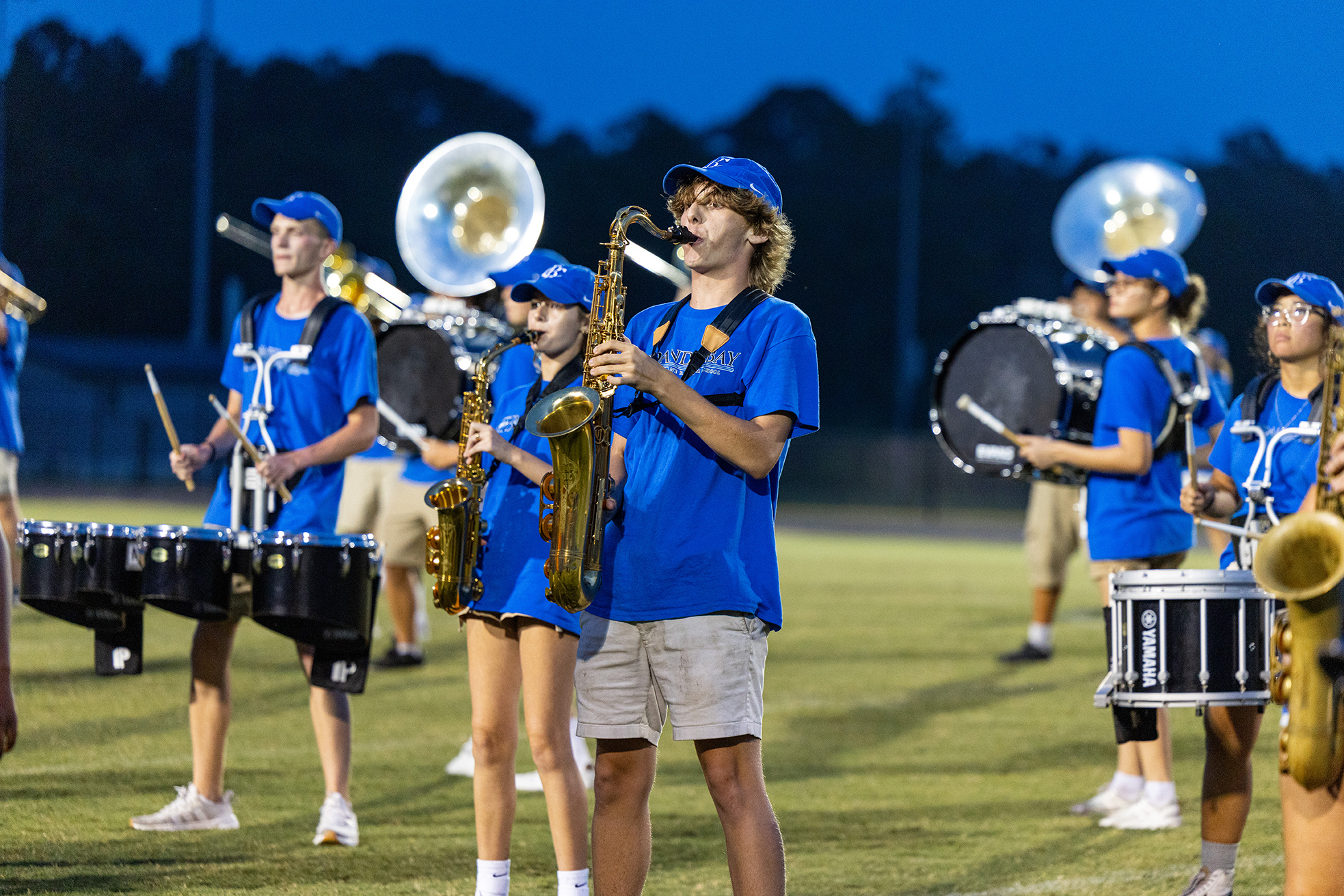 A band on a grassy field plays instruments at night. Most wear blue shirts and khaki shorts.