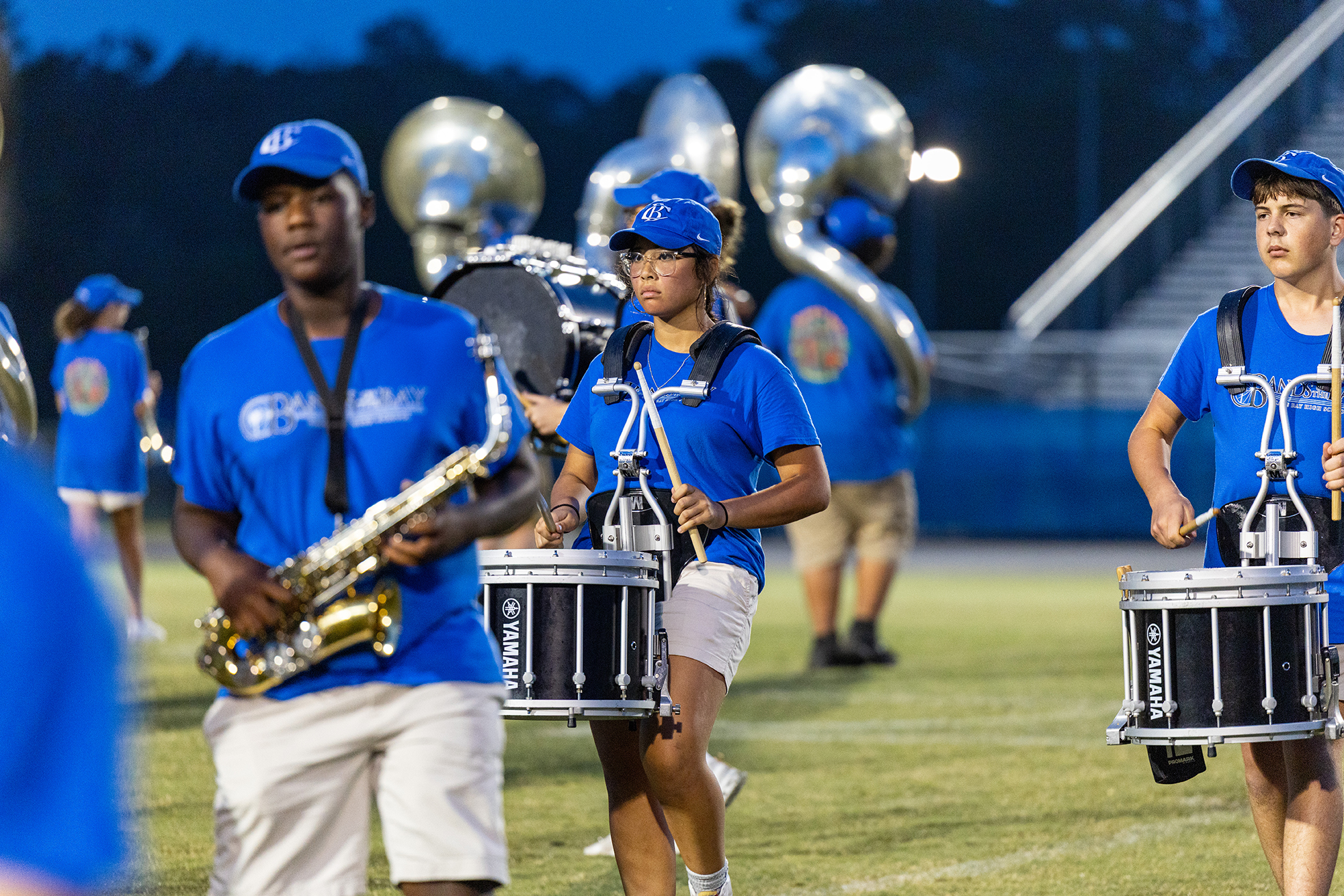 Several young people play instruments on a football field, including drums, a saxophone, and large brass instruments.