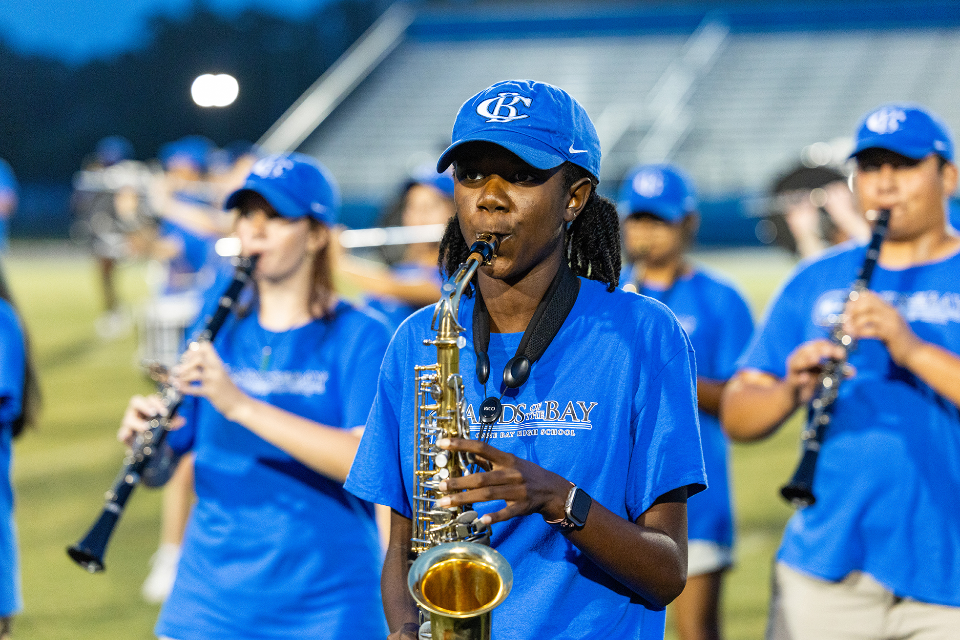 Band members in blue uniforms and blue caps play musical instruments on a grassy field.