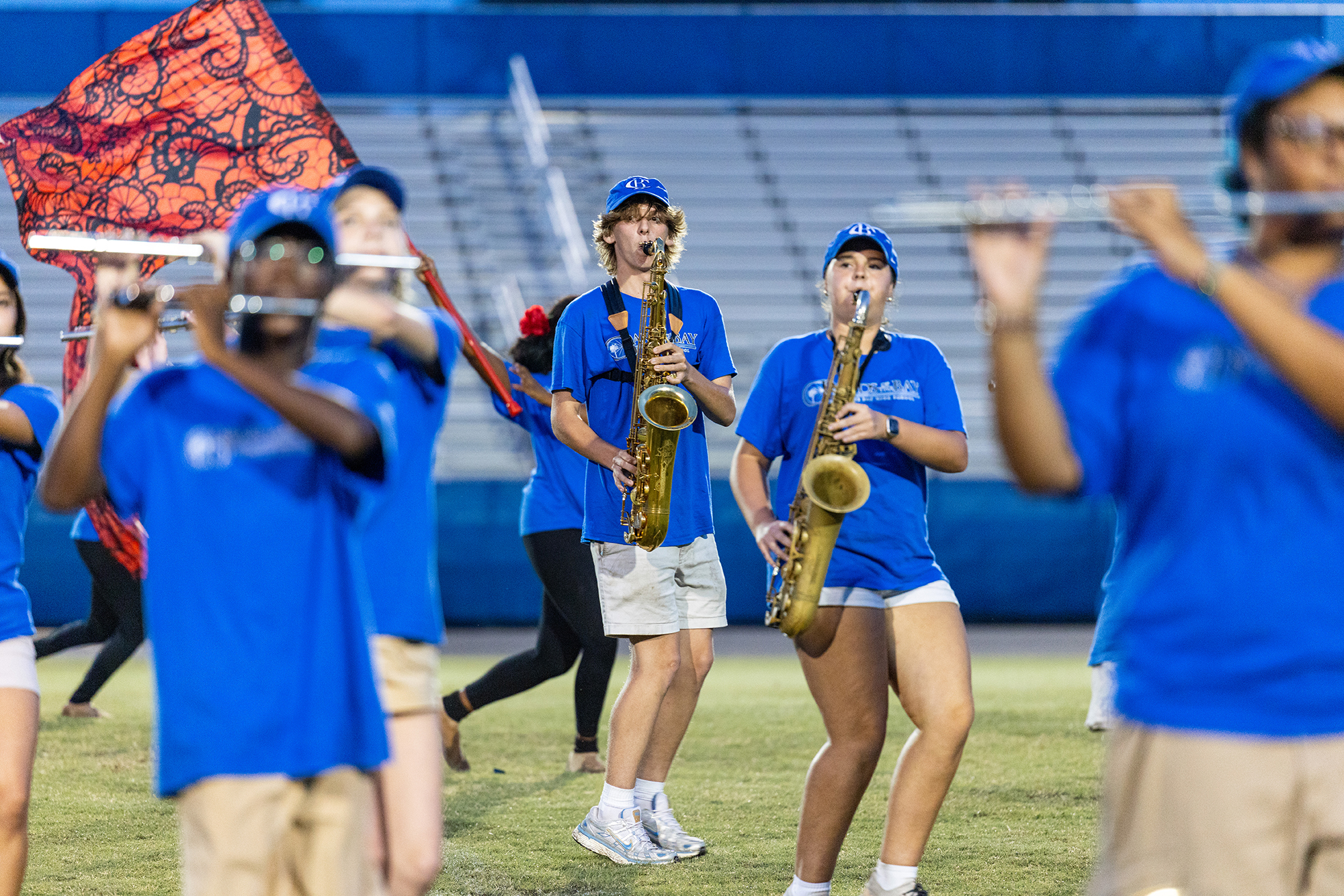A band with blue uniforms and caps plays instruments on a grassy field. Some play flutes, others saxophones.