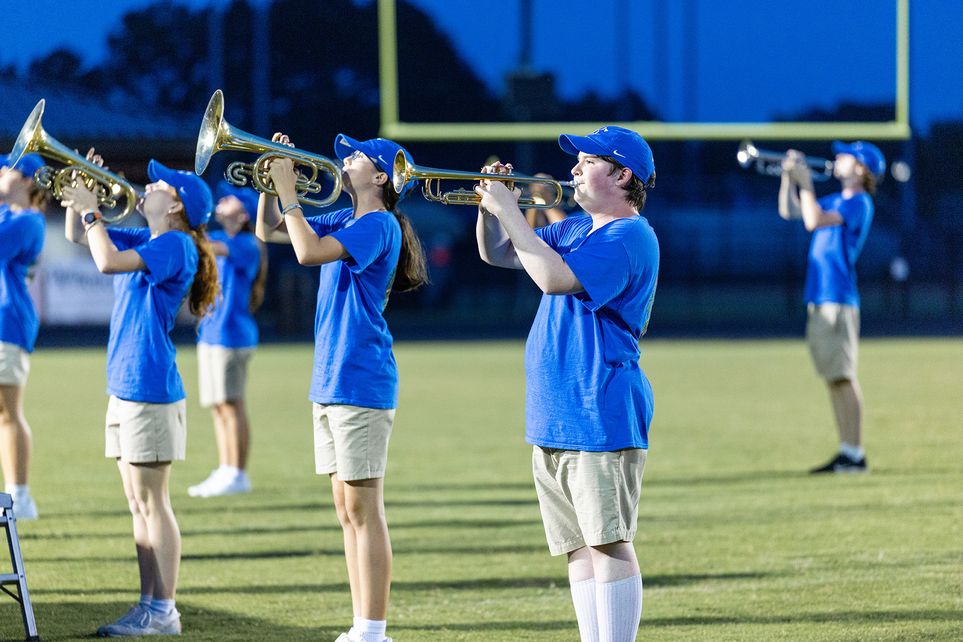Six people in blue shirts and shorts play trumpets on a football field at night.