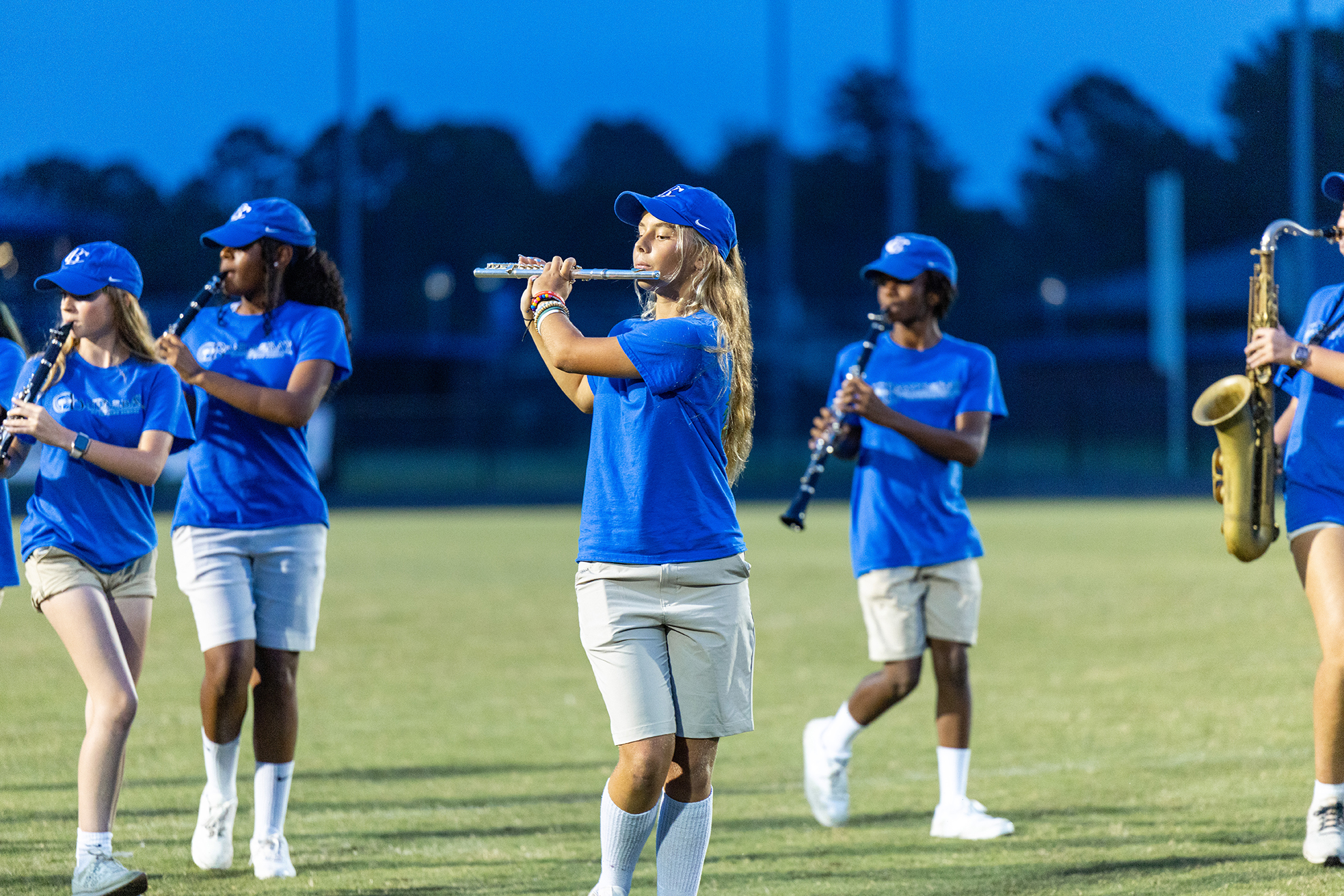 Four individuals in blue shirts and white shorts perform on a field. One plays the flute, another the saxophone, and two play clarinets.