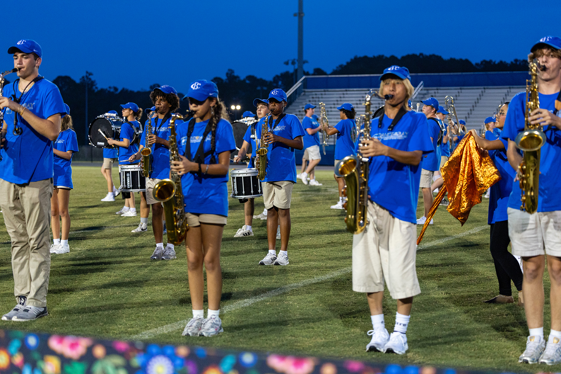 A marching band in blue uniforms performs on a field at night, with a stadium in the background.