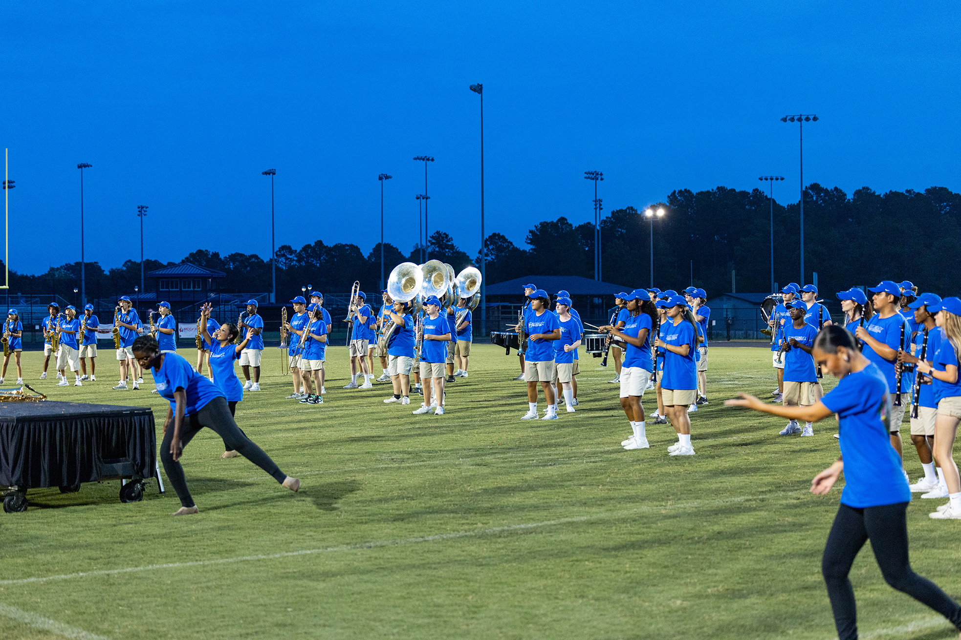 A group of people in blue uniforms practice on a field at dusk, some holding musical instruments.