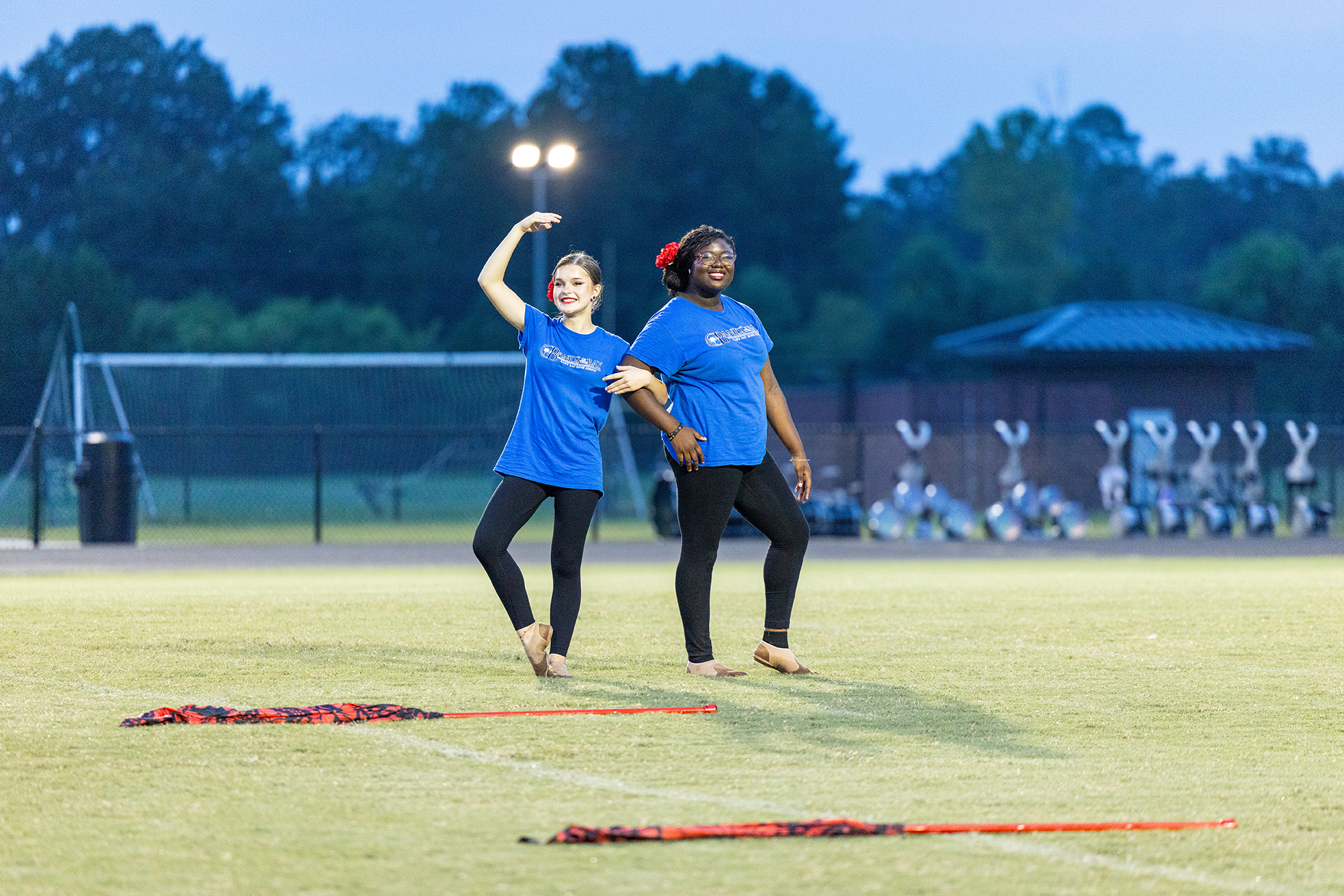 Two women dance in blue shirts and black pants on a field. One holds a hand up.