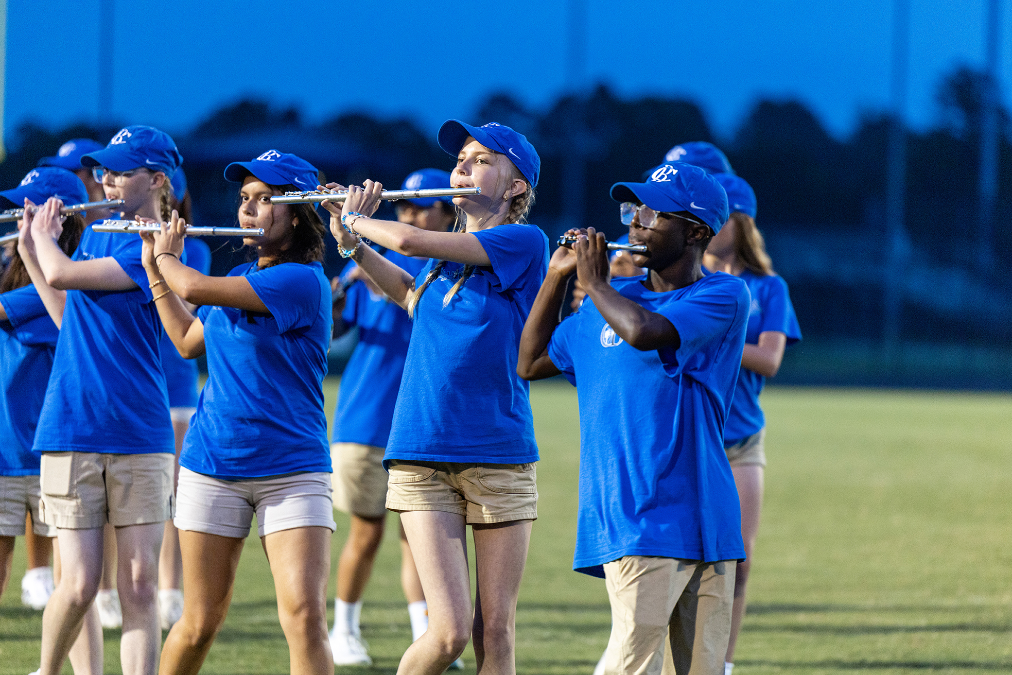 Group of individuals in blue shirts and white shorts playing flutes during a performance on a field.
