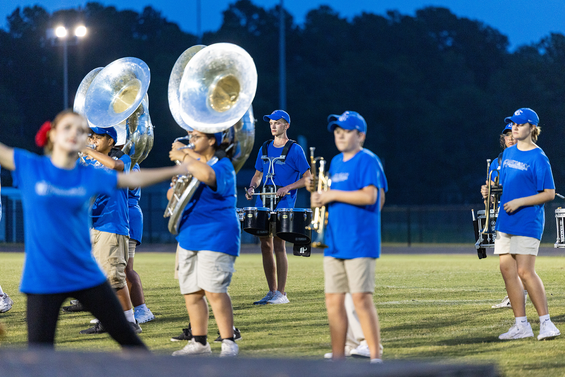A group of people in blue shirts and white shorts perform on a field, playing large instruments.