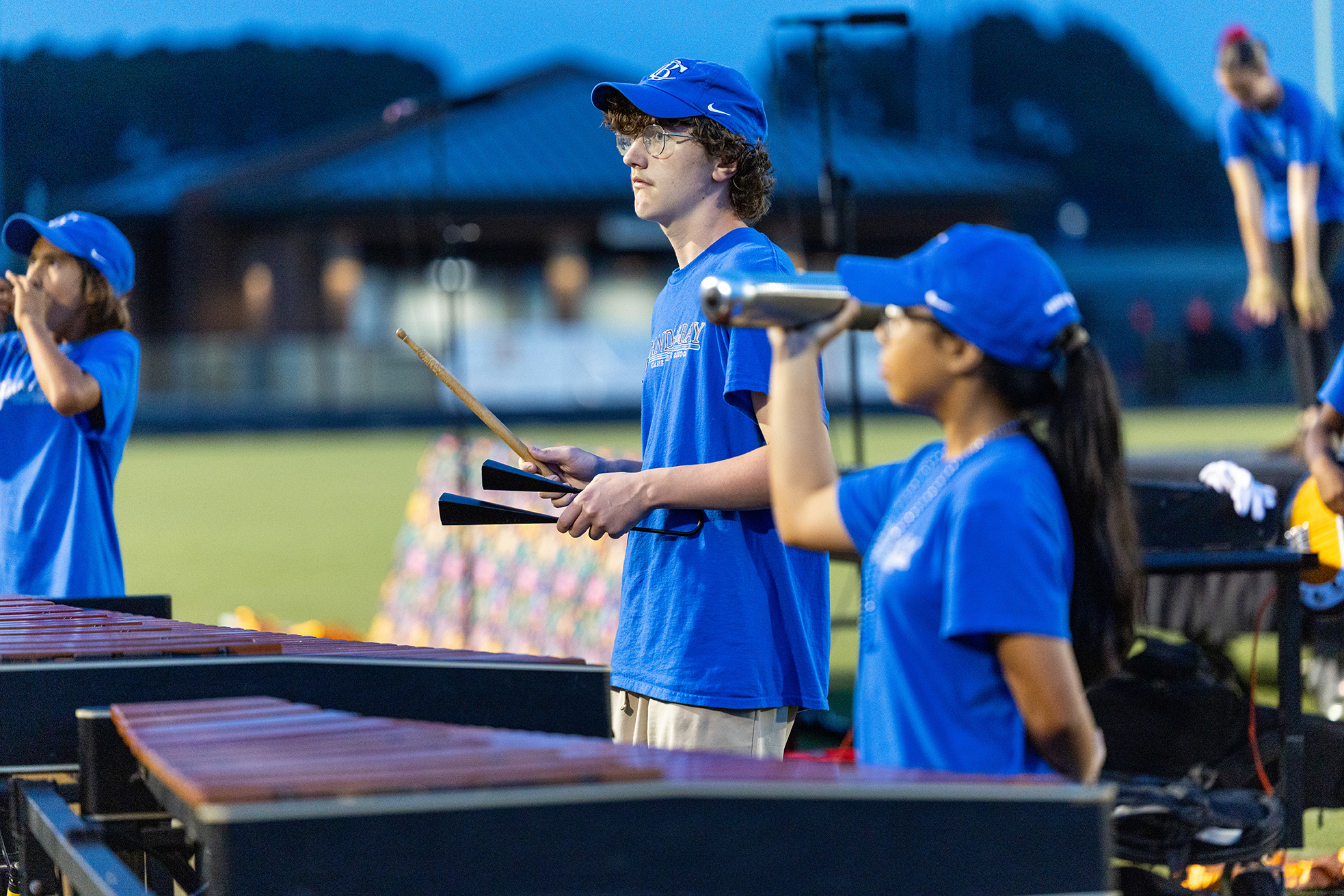 Four people in blue shirts play instruments on a field. One plays a marimba, another plays xylophone.