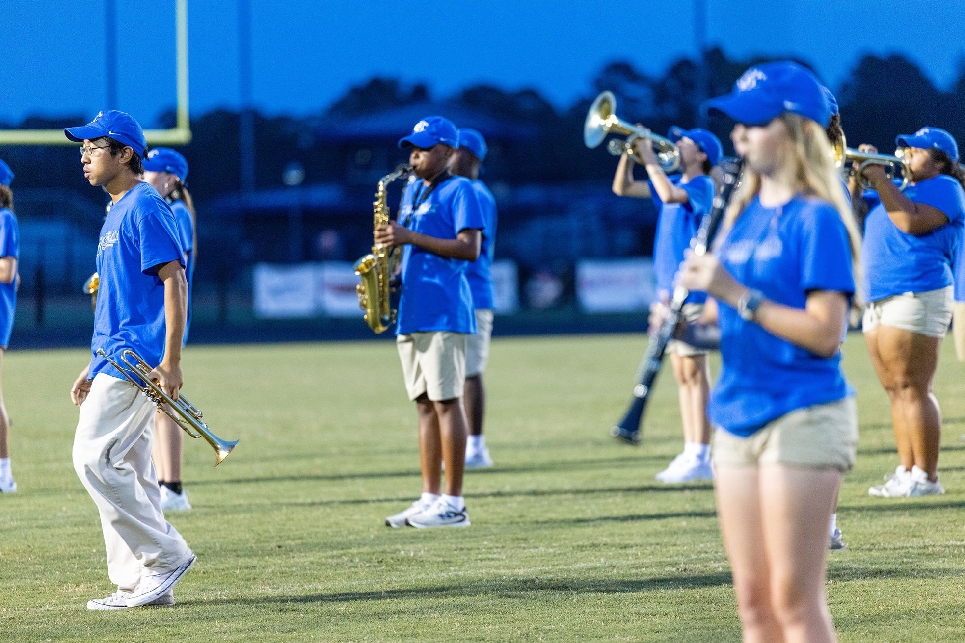 A group of students in blue uniforms stands on a field, playing trumpets, saxophones, and other instruments.