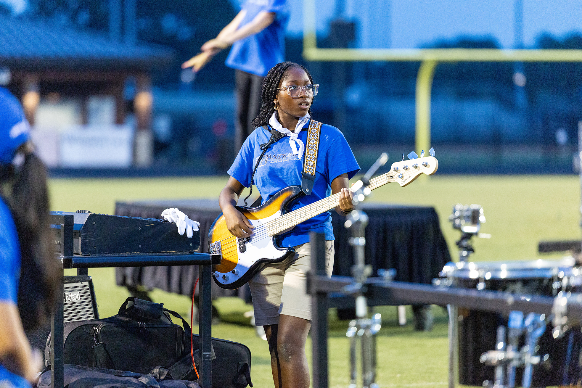 A woman in a blue shirt plays an electric guitar on a stage, with a black backdrop.