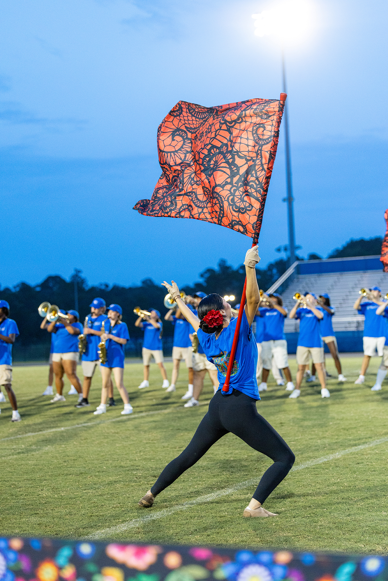 Person in blue shirt and black pants holding a red flag, standing on a football field with band members behind.