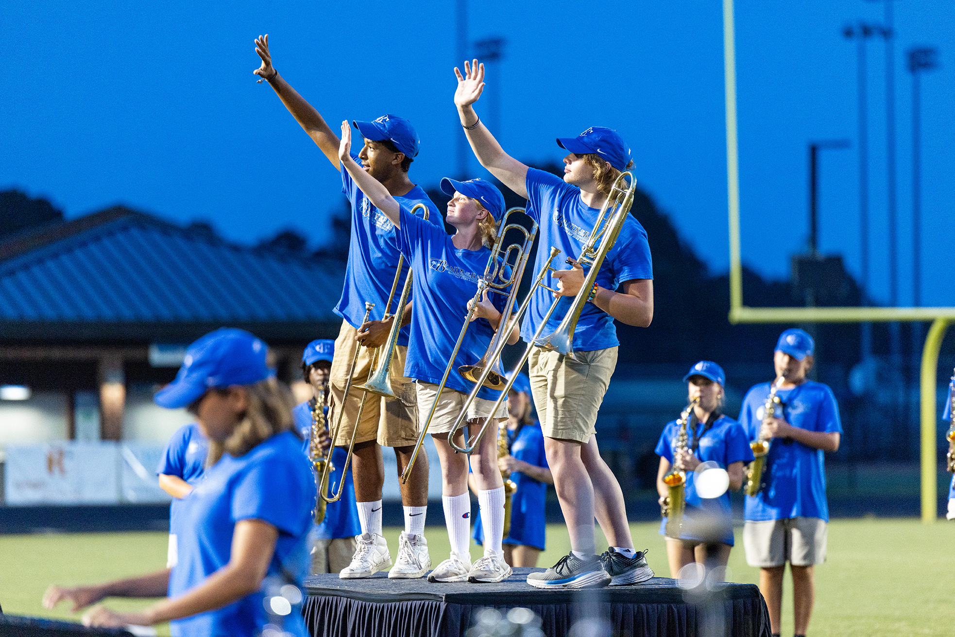 A marching band performs on stage with instruments. They wear matching blue shirts and caps. The field has a goal post.