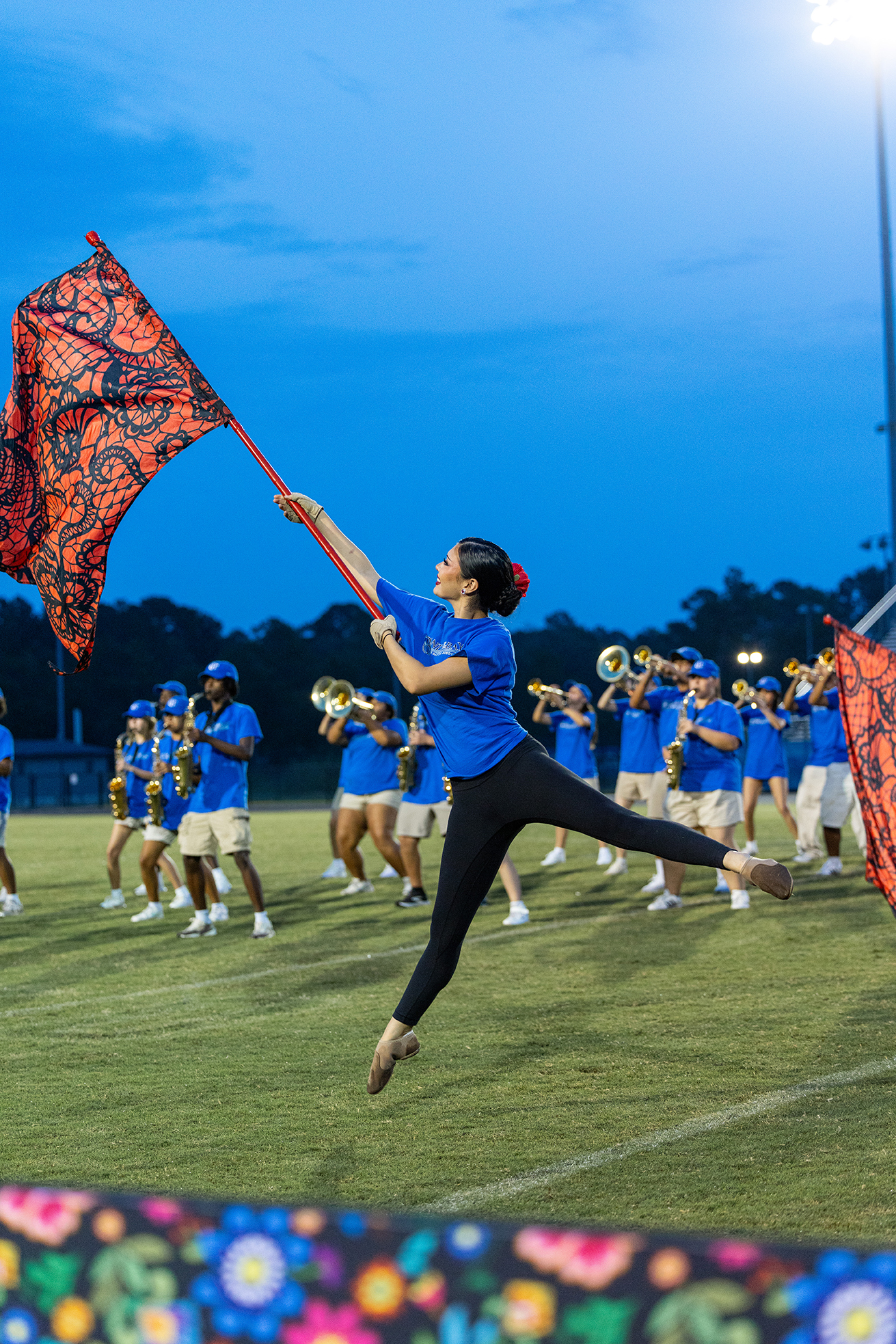 A woman in a blue shirt and black leggings jumps, holding a red flag. Behind her, a marching band practices on a grassy field.