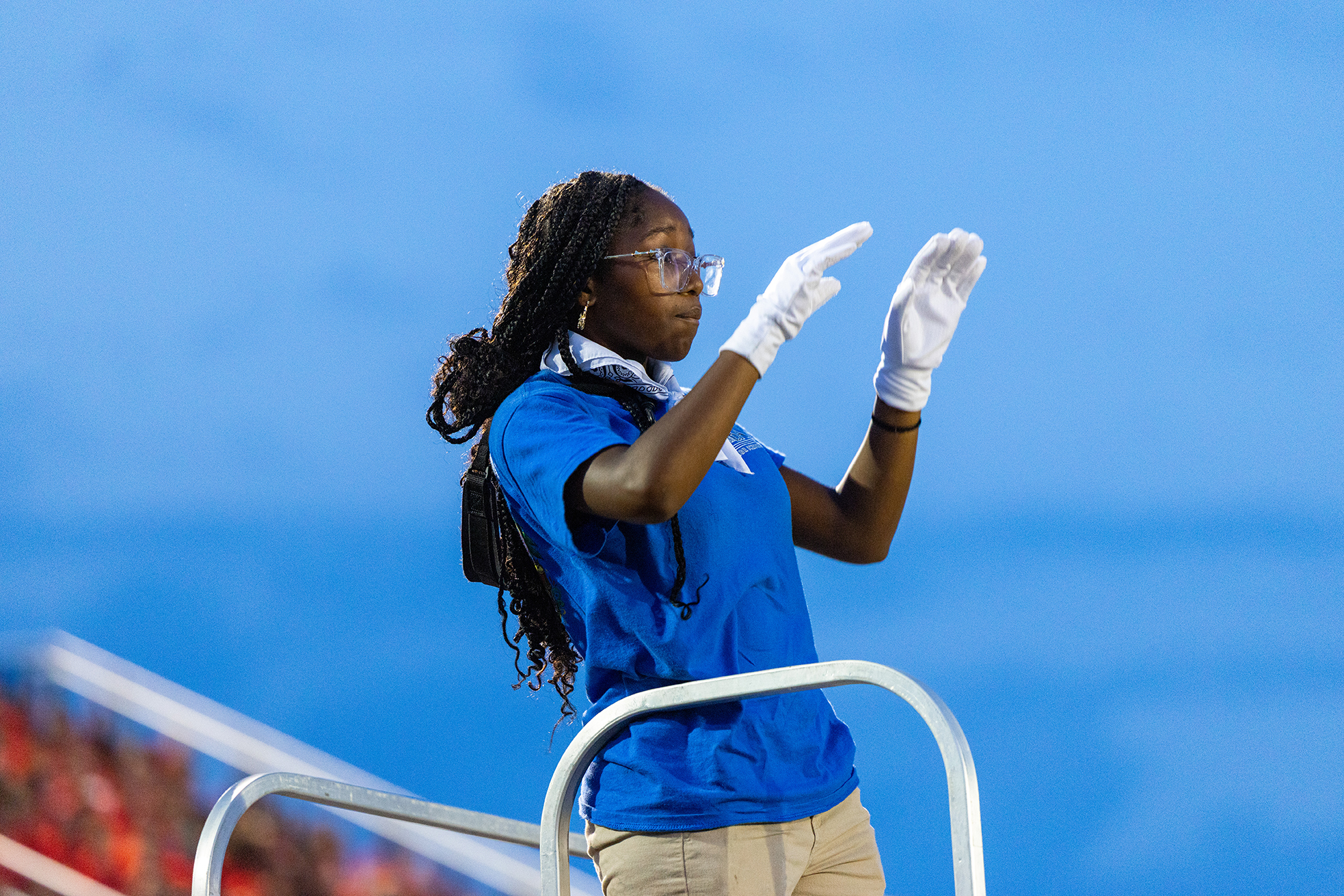 A woman in a blue shirt, white gloves, and glasses stands on a platform, gesturing upwards.
