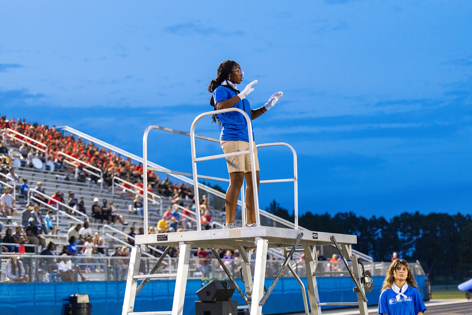 Person in blue shirt, white gloves, and skirt stands on a metal platform over an audience.