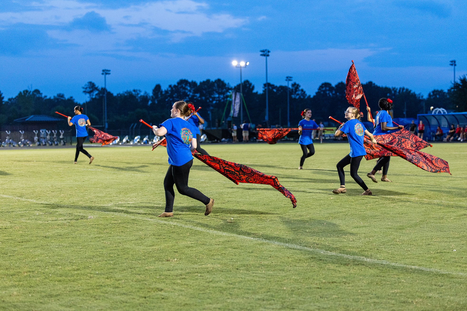 A group of people on a field perform a synchronized flag routine with orange flags, wearing blue shirts and black pants.