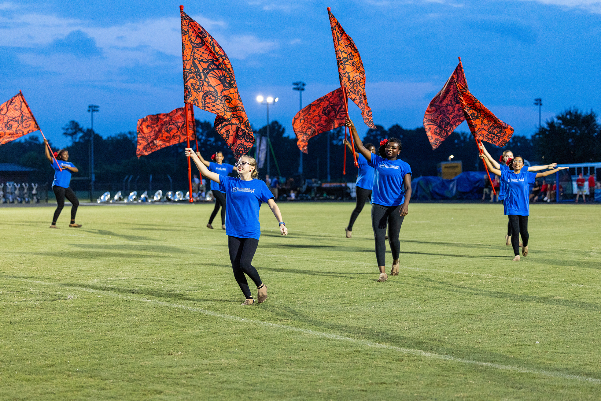 Five people in blue shirts, black pants, and sneakers wave orange flags on a football field.