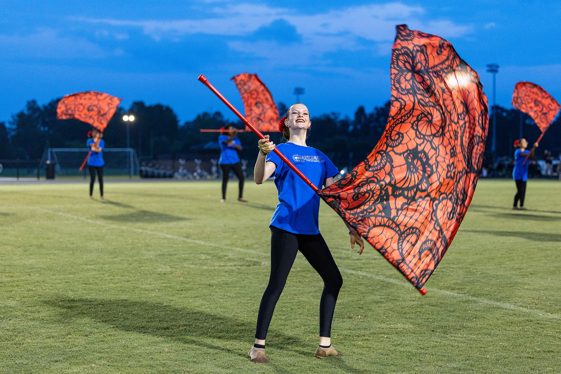 A woman in blue holds an orange flag while standing on a field, with other performers in similar poses.