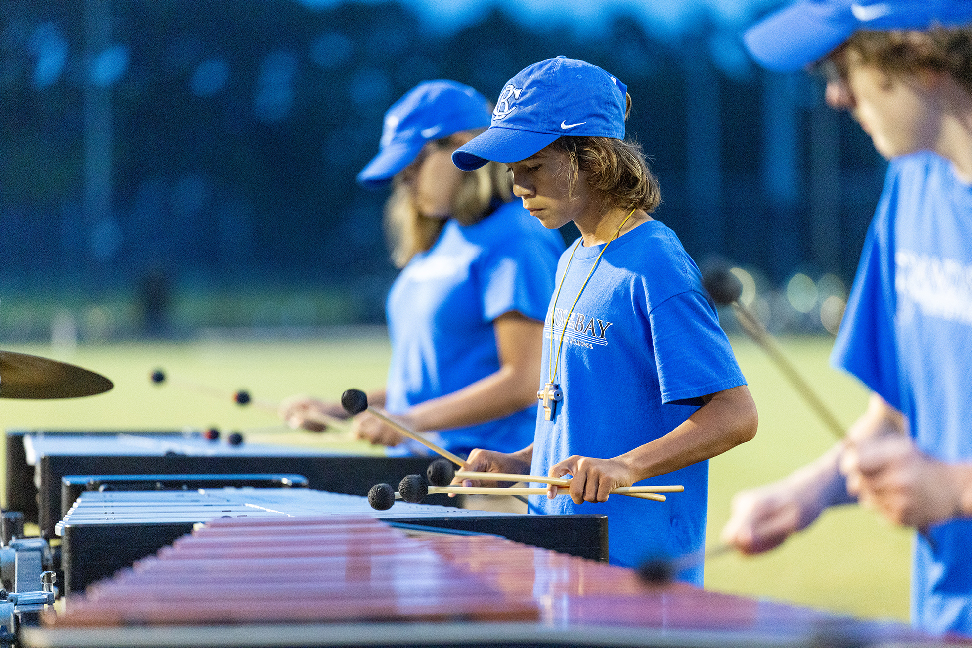 Four individuals in blue shirts and caps play xylophones, with blurred trees and a street light in the background.