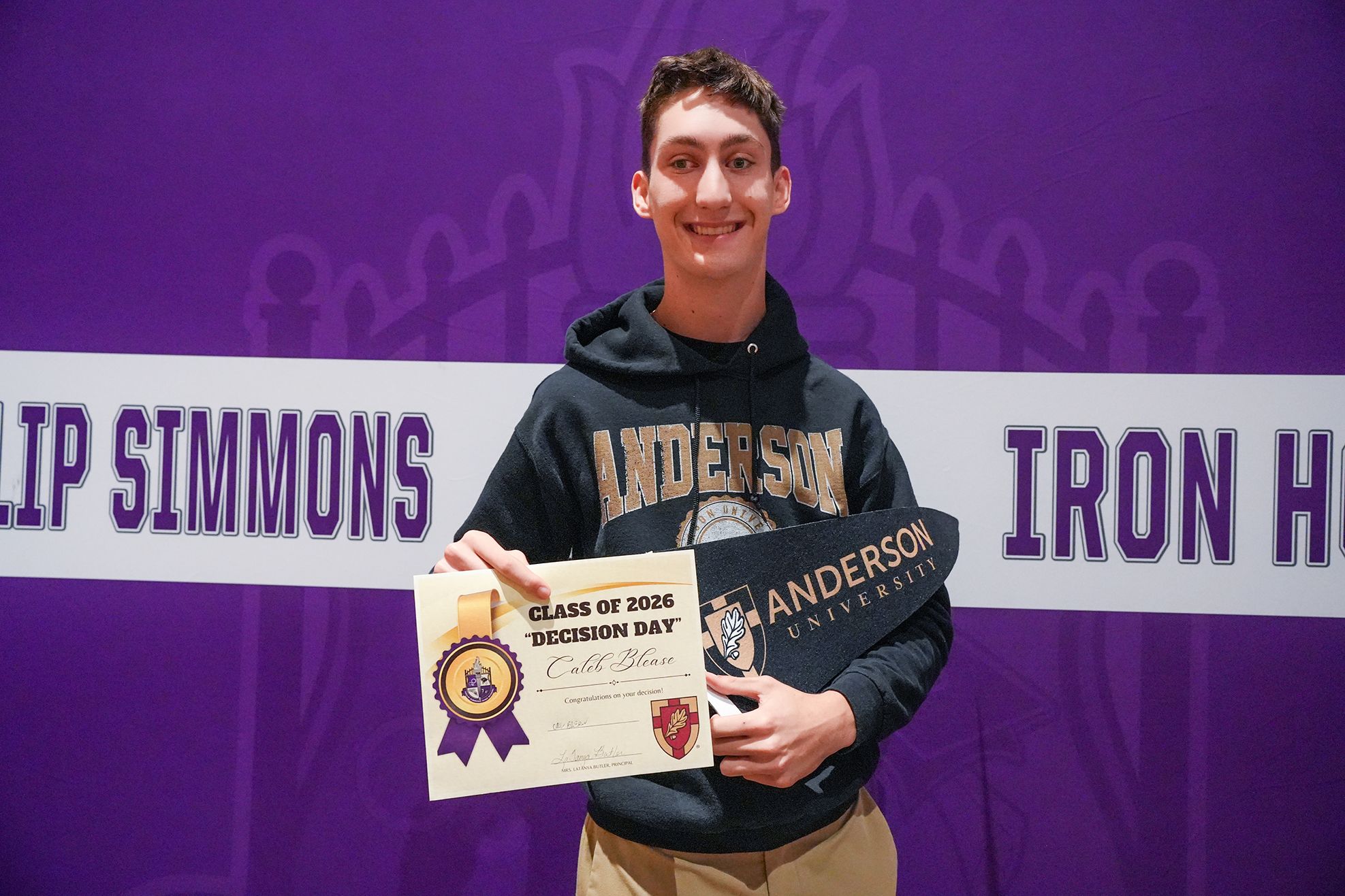 Person in black hoodie holds a certificate against a purple backdrop with "Philip Simmons" and "Iron Hill" banners.