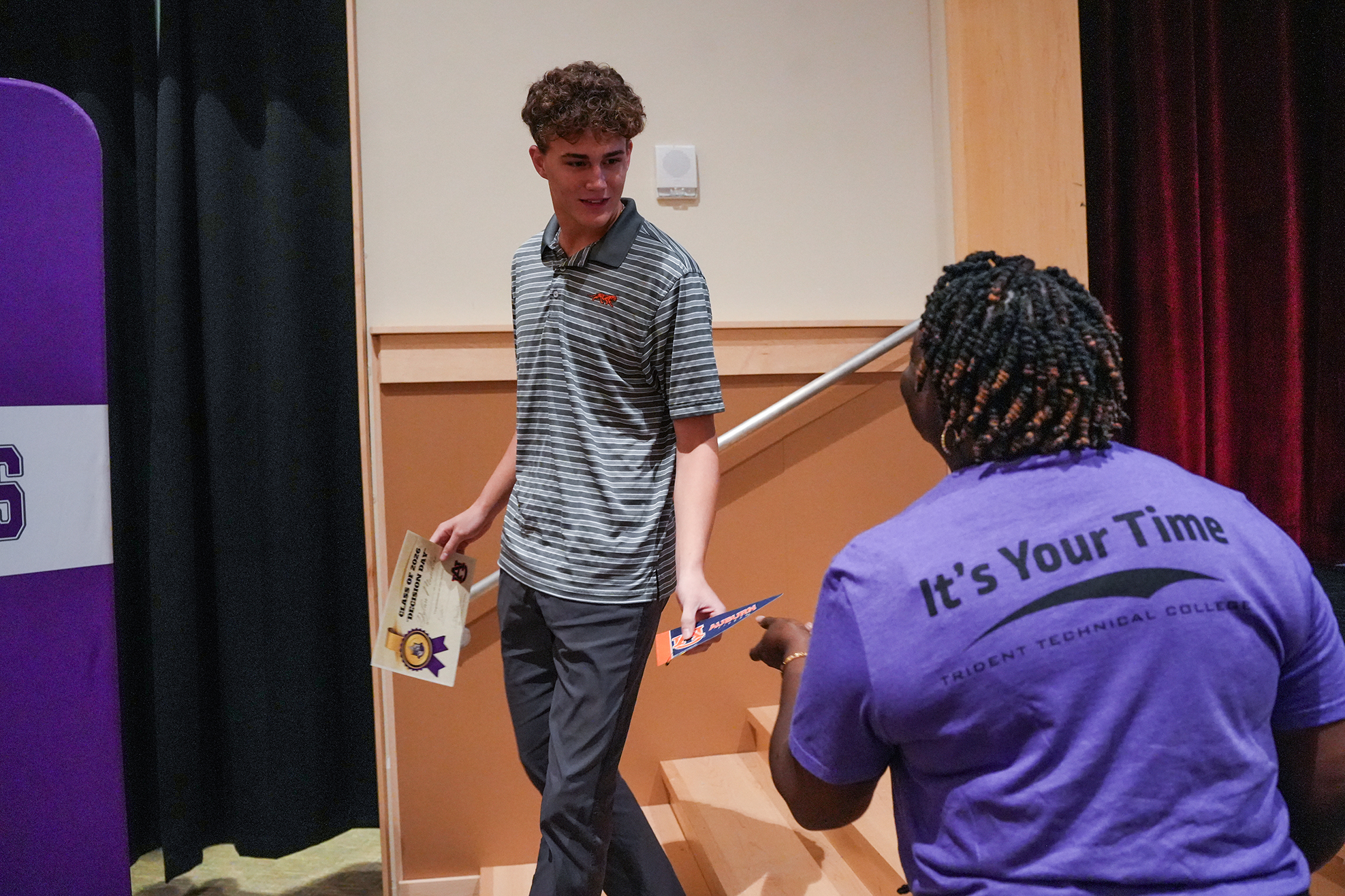 A man in a striped shirt walks up steps with papers in hand, another woman in a purple shirt hands him something.