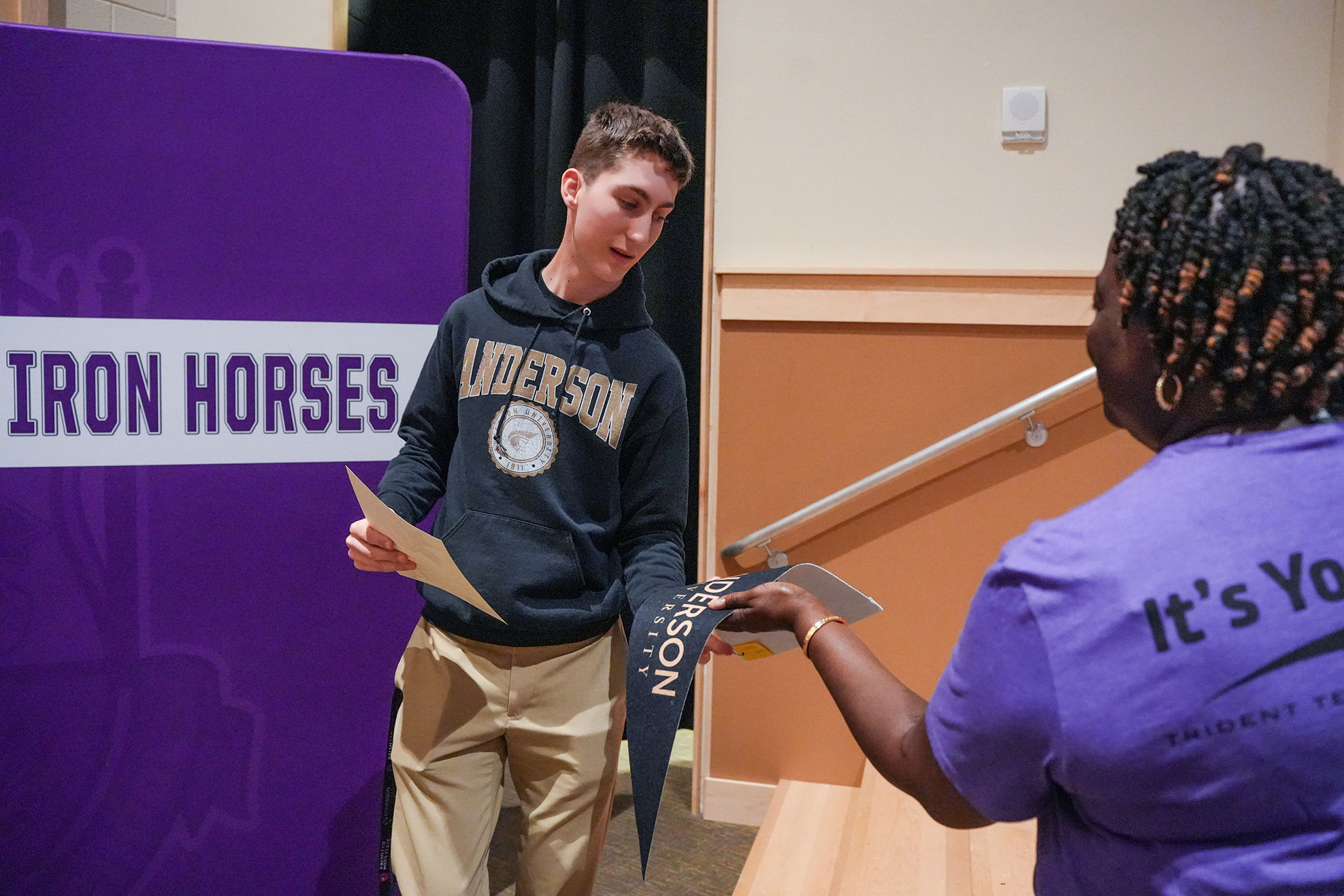 A boy in a black hoodie and khaki pants holds a banner. A woman in a purple shirt presents him with a certificate.